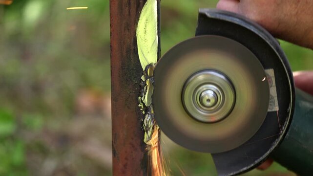 Metal worker uses electric angle grinder to polish welded seam on steel post, bright orange sparks flying as abrasive disc smooths rusty metal surface outside on sunny day