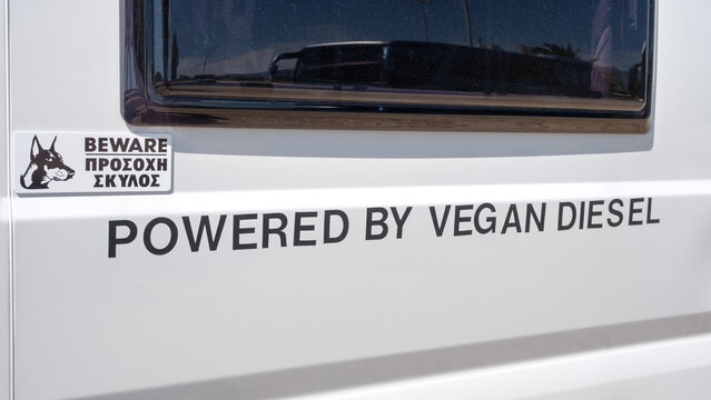 Close-up of white vehicle featuring 'Powered by Vegan Diesel' decal and bilingual warning sign in front of dog, Corinth, Greece.