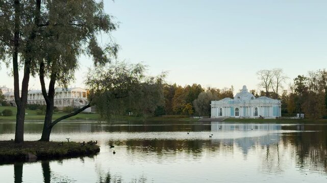 A pavilion stands beside a calm lake reflecting tall trees. The serene landscape creates a peaceful atmosphere. This scenic view is in Catherine Park, Pushkin, Saint Petersburg.