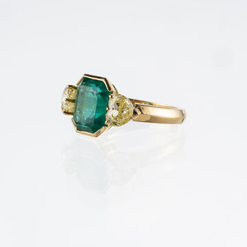 18kt yellow gold ring with a central emerald and two heart-cut diamonds. Photographed on a white background.