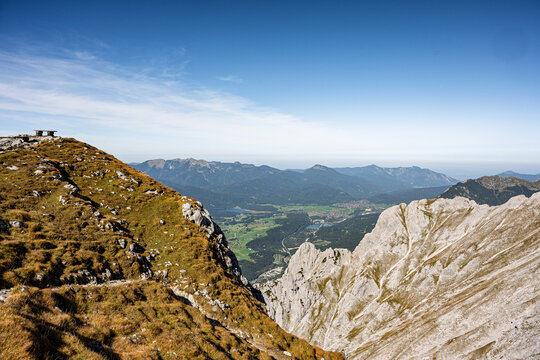 High angle panoramic view from the rocky peaks of the Karwendel Alps featuring a wooden bench on a grassy ridge overlooking the valley below.
