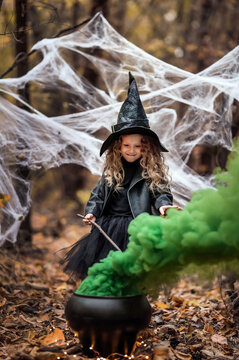 Little girl in a witch outfit holding a magic stick over a cauldron with thick green smoke