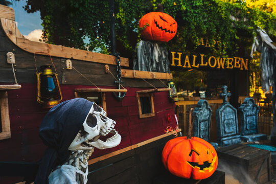 Halloween pirate ship decoration background with skeleton pirate with bandana, carved jack o lantern pumpkins, gravestones, and Happy Halloween sign outdoor. Spooky Halloween party outdoor decoration.