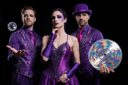 Three circus performers wear purple costumes in studio. Male juggler holds magic crystal ball woman stands in center. Man holds large holographic disco disc. Pro entertainers pose with props against