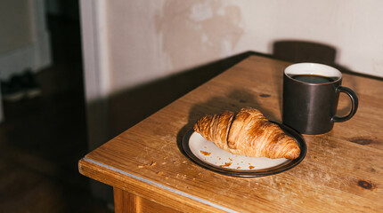 Fototapeta premium Fresh croissant on a plate with a cup of coffee on a rustic wooden table, cozy breakfast scene with warm light and copy space