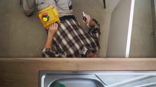 Top-down view of a skilled handyman lying on the kitchen floor, using an adjustable wrench to fix a pipe underneath the sink during a routine home maintenance service call