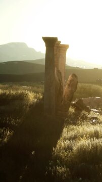 Two stone pillars stand in a grassy area during sunset. Shadows stretch across the ground as the sun dips behind distant hills, creating a warm atmosphere around the scene.