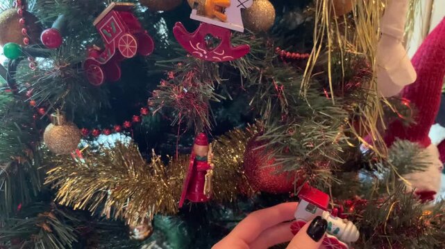 Close-up view of a hand with dark nails hanging traditional red wooden ornaments and golden tinsel on a decorated evergreen Christmas tree.   