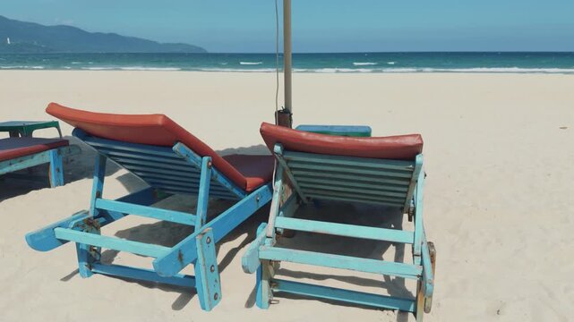 Da nang city. Weathered blue beach chairs with orange cushions stand beneath beach umbrella, overlooking gentle waves. Empty seaside seating offers peaceful place to relax beside distant mountains.