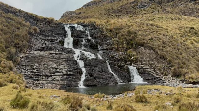 Water cascades down dark stepped rock face into clear pool surrounded by yellow grass in viconga valley during cloudy day in cordillera huayhuash peruvian andes