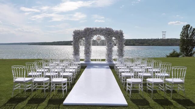 An elegant floral wedding arch stands on a grassy lawn overlooking a calm lake with rows of clear chairs arranged for an outdoor marriage ceremony