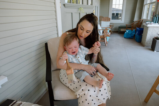 Mother comforting crying toddler on porch holding paintbrush