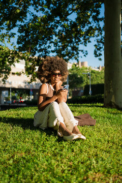 Woman with Afro Hair Sitting on Park Grass Using Smartphone