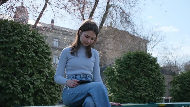 Concentrated young woman sitting on a park bench during spring, deeply engaged in reading an ebook on her electronic device, preparing for exams or enjoying a digital novel outdoors