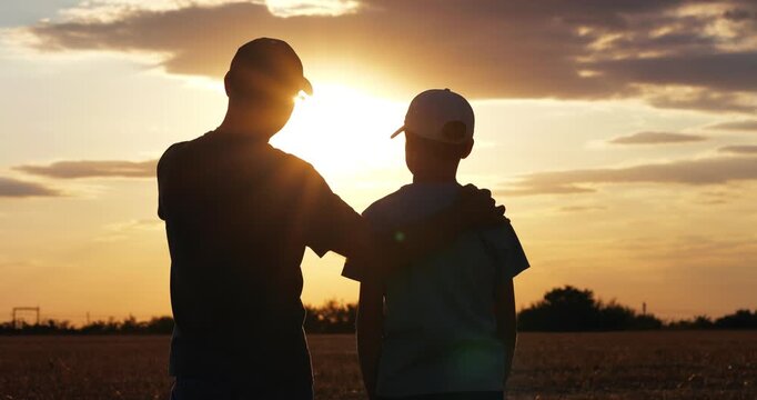 Father and son stand together in field at sunset. Silhouetted family shares quiet moment across rural landscape. Golden light evokes connection, love, nature, and peaceful outdoor living.