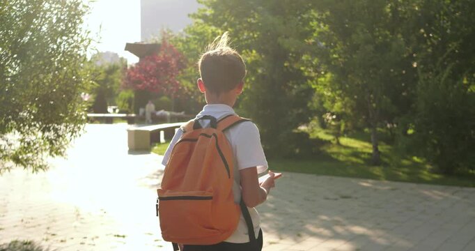 Schoolboy with orange backpack walks through sunny park, stops at pedestrian crossing paths and checks smartphone for navigation. Child chooses route and turns onto another path in urban surroundings