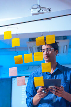 Smart developer holding tablet and reviewing project structure on glass board with sticky notes in tech office.