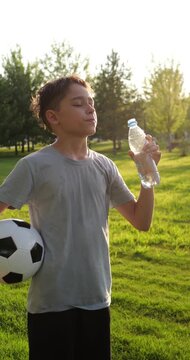 Boy drinks water from bottle after soccer practice in sunny park. Young athlete cools down on green grass while holding football. Healthy outdoor lifestyle shows hydration, sport, and summer wellness.
