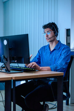 Male customer support representative wearing headset working on computer in office night shift, professional call center agent talking online and typing on keyboard