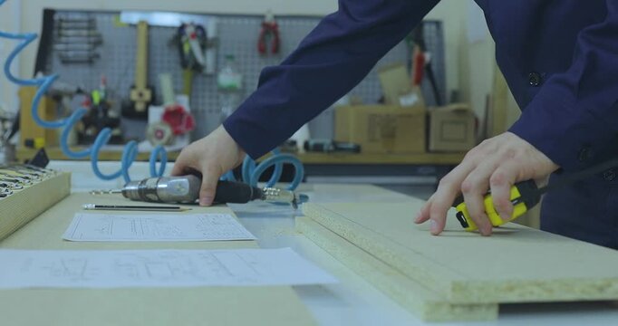 A furniture maker in blue workwear measuring a particle board part with a tape measure on a workbench in a manufacturing workshop. Close-up