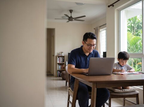 Malaysian Father Working From Home With Child - Malaysian father working on a laptop while his child draws nearby, with copy space for work-life balance and family advertising.