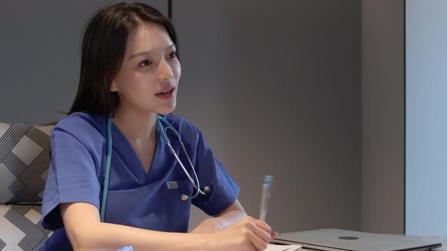 Asian female surgeon in blue scrubs talks with patient to ask about symptoms, takes a note on document clipboard in clinic before examining. Beautiful woman doctor put on rubber gloves before check-up