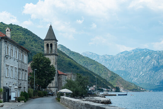 Coastal road winds past historic stone buildings and a church bell tower in Stoliv, Montenegro, along the Bay of Kotor