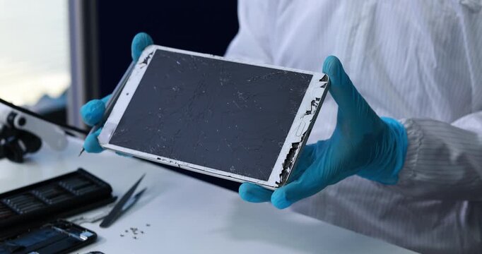 Technician in blue gloves holds a damaged tablet with cracked screen and broken edges during electronic device repair at a workbench.