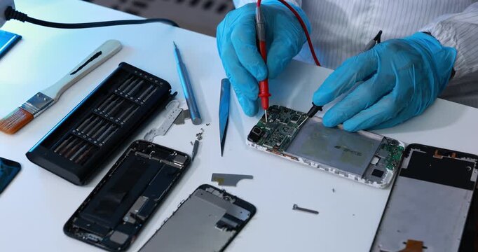 Technician wearing blue gloves tests an opened smartphone circuit board with multimeter probes beside repair tools and removed phone parts on a workbench.