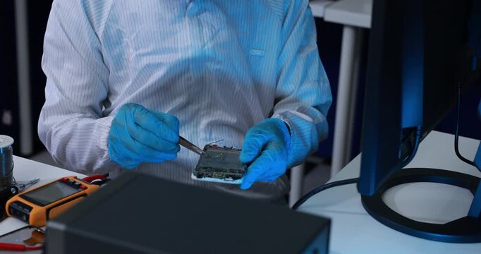 Technician in blue gloves examines a circuit board with tweezers at a laboratory workstation beside a computer monitor and measuring device.
