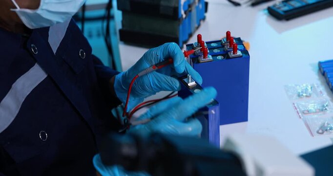 Technician wearing blue gloves tests battery cells with probes at an electronics workbench, checking voltage during assembly or repair.