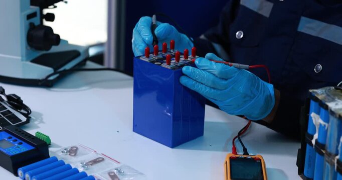 Technician wearing blue gloves checks a rechargeable battery pack with test probes at an electronics workbench, repair, maintenance and energy storage work.