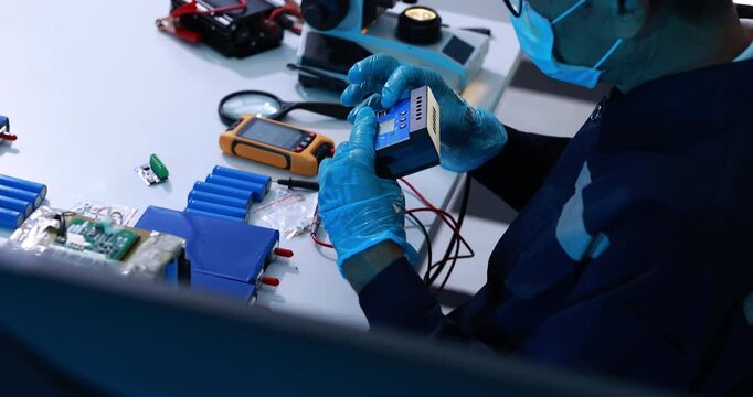 Electronics technician wearing gloves tests a small device with meter probes at a workbench filled with circuit boards, batteries and repair tools.