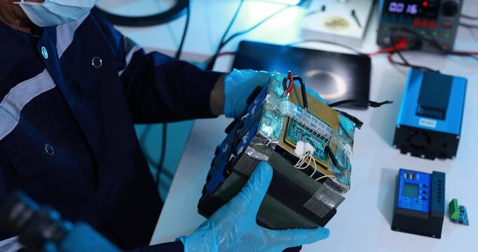 Close-up of a technician wearing gloves and a face mask inspecting a damaged electronic device with exposed circuit board and wires at a repair workstation.