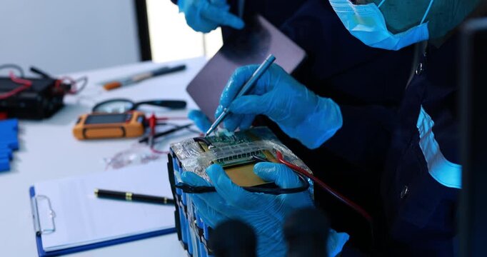 Technician wearing blue gloves repairs an electronic battery pack with probes and tools at a workbench in a modern workshop.