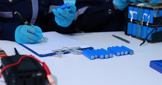 Forensic investigators in blue gloves document evidence beside batteries, test equipment and wires on a laboratory table.