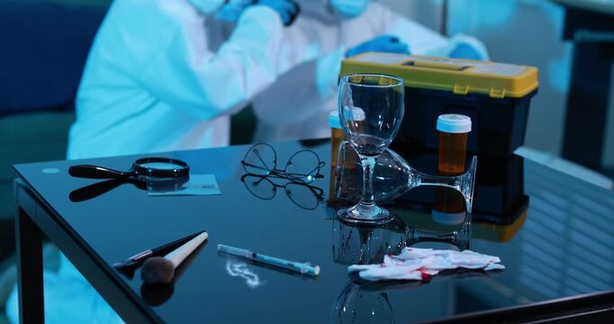 Forensic evidence on a reflective table at a crime scene, including a wine glass, pill bottle, syringe, gloves, scissors and a yellow case while investigators work in the background.