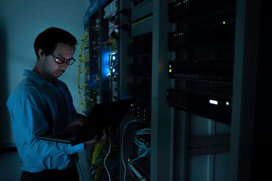Focused Asian male IT network administrator uses a laptop to monitor server performance inside a dark data center room managing complex corporate cybersecurity operations and infrastructure.
