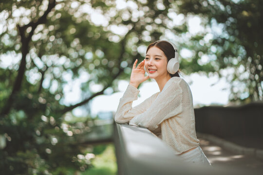 Happy young Asian woman wearing wireless headphones outdoors, smiling while touching earphone and enjoying audio during relaxing leisure time in green park.