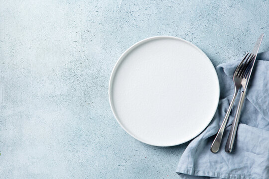Empty white plate with knife and fork on a light blue concrete background. Copy space, close up, top view.