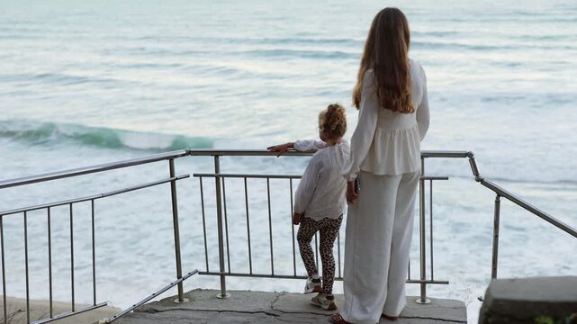 Back view of young mother standing next to her cute little daughter looking together at blue ocean waves from viewing deck railing during seaside vacation