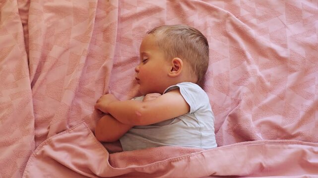 Cute toddler boy sleeping peacefully in bed under soft pink blanket during daytime nap at cozy home bedroom childhood rest relaxation concept