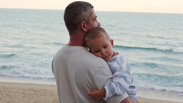 Tired toddler boy leaning on his father shoulder while standing on seaside beach against sea waves during family vacation travel