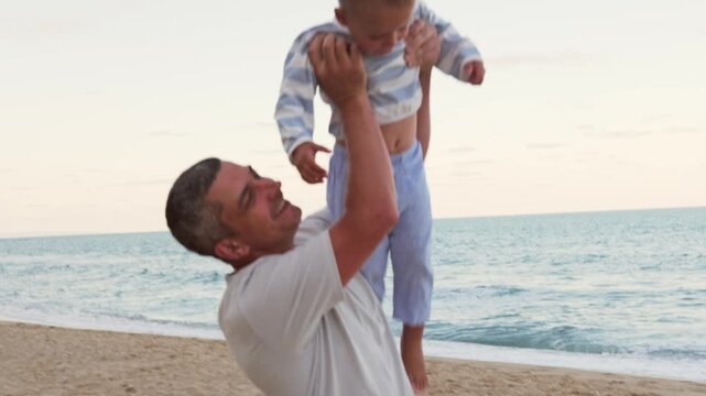 Happy laughing father lifting his joyful little toddler son in arms on sandy beach near sea having fun together