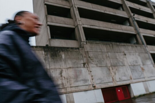 Brutalist concrete housing block with a woman in a dark rain jacket walking across a wet, grimey forecourt for urban lifestyle