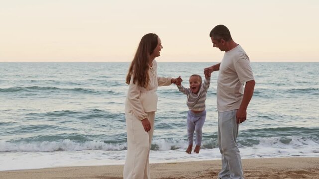 Happy family with toddler son having fun and playing together on sandy sea beach during evening summer vacation