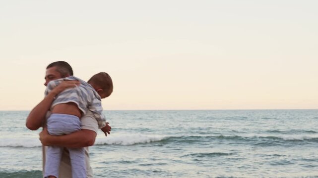 Side view of happy father holding and lifting his cute toddler son in air near sea shore on sandy beach