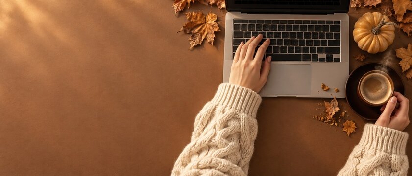 Hands in cozy cable knit sleeves using a laptop on a brown desk with pumpkins, autumn leaves, and steaming coffee for home office and blog mockup