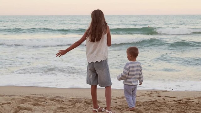 Older sister and little brother standing on sandy sea beach shore and looking at ocean waves during sunset