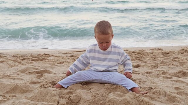 Cute little boy sitting on sandy sea beach and playing with sand during peaceful family summer vacation